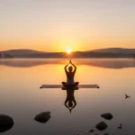 Serene sunrise over lake with person meditating, embodying mindful morning habits.